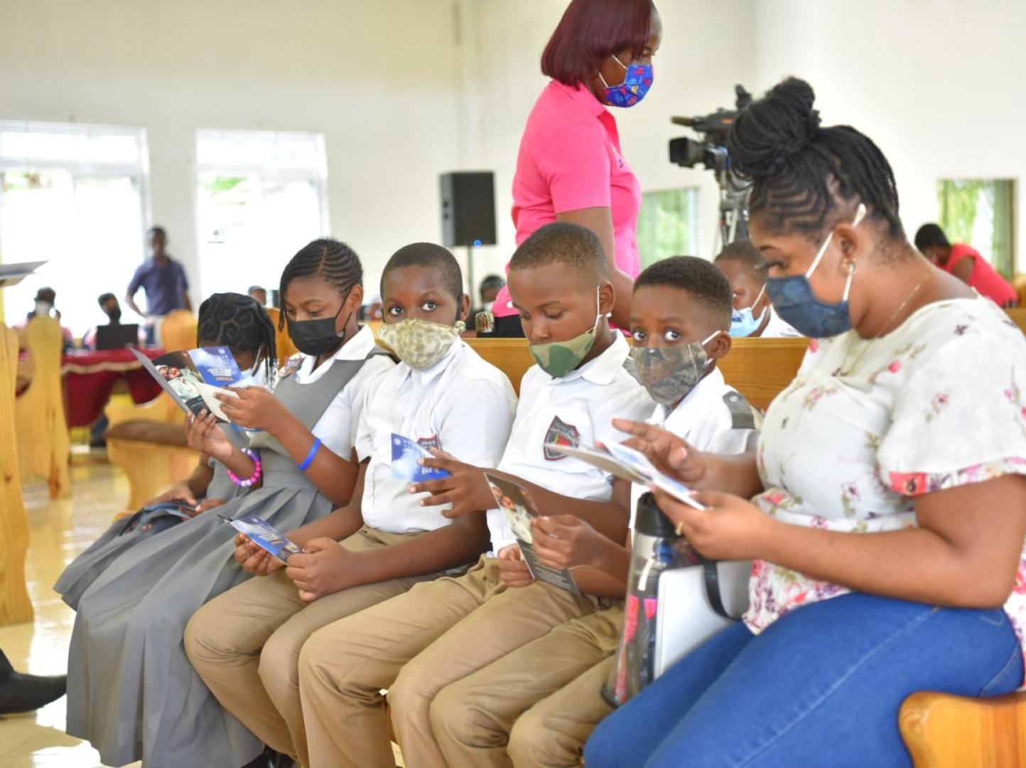 Young students seated with masks on with their teacher on a bench reading books.