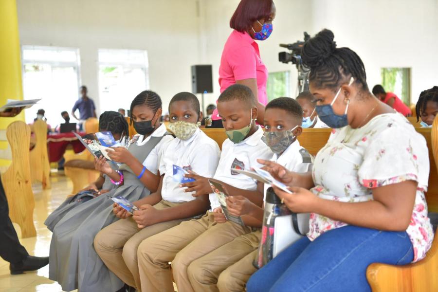 Young students seated with masks on with their teacher on a bench reading books.
